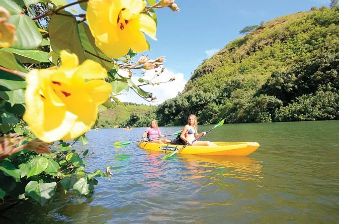 Two women in swimsuits paddling a yellow tandem kayak past yellow hibiscus flowers